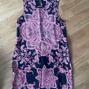 Mönstrad kortklänning från Indiska - Supersnygg kortklänning från Indiska med ett stort paisleyinspirerat mönster i rosa och lila på marinblå botten. Ärmlös modell med rund halsringning, perfekt för dig som gillar färg och mönster. Lätt och skönt material som känns luftigt. S-M