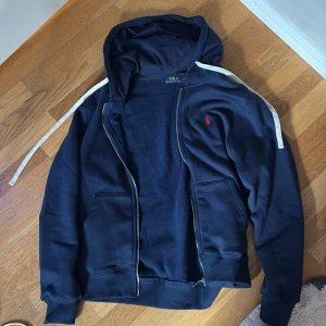 Marinblå hoodie från Polo Ralph Lauren - Snygg marinblå hoodie från Polo Ralph Lauren med dragkedja, huva och vita snören. Klassisk röd logga broderad på bröstet och två fickor framtill. Perfekt för en chill och stilren look.