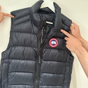 Svart dunväst från Canada Goose - Säljer en svart dunväst från Canada Goose med klassisk logga på bröstet. Västen har hög krage, quiltad design och dragkedja framtill. Perfekt för lager-på-lager och riktigt snygg till streetwear eller chillare outfits.