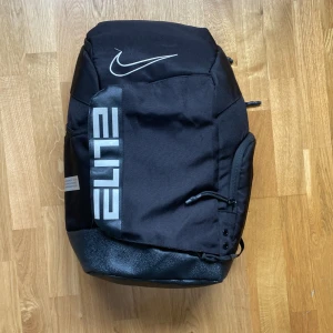 Nike Elite svart ryggsäck - Snygg svart Nike Elite ryggsäck med stora loggor och reflekterande detaljer på axelremmarna. Rymligt huvudfack, flera dragkedjefickor och slitstark botten. Perfekt för att bära träningskläder eller skolgrejer. Justerbara remmar för bästa passform.