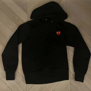 Svart Comme des Garçons Play hoodie - Svart hoodie från Comme des Garçons Play med klassisk huva och snörning. På bröstet sitter det ikoniska röda hjärtat med ögon. Passar folk som är runt 164, både män och kvinnor. Är storlek M men är väldigt liten.