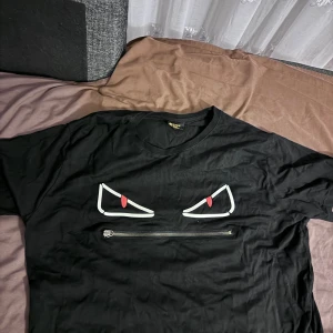 Svart Fendi t-shirt med ögon och dragkedja - Svart t-shirt från Fendi med coolt tryck av vita och röda ögon på bröstet och en silvrig dragkedja under. Klassisk rund halsringning och korta ärmar. Tillverkad i mjuk bomull, perfekt för dig som vill sticka ut med en unik designerpiece.
