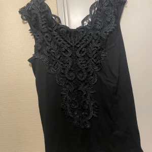 Svart topp med spetsdetaljer - Svart ärmlös topp med snygga spetsdetaljer och broderat mönster framtill. Toppen har rundad halsringning och är i en rak, kort modell. Perfekt för dig som gillar stilrena plagg med en twist.