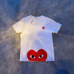 Vit Comme des Garçons Play t-shirt - Vit t-shirt från Comme des Garçons Play med ikoniskt rött hjärta med ögon på bröstet och ett stort hjärta med ögon tryckt längst ner. Klassisk rund hals och korta ärmar. Tillverkad i mjuk bomull, perfekt för en clean och cool look.
