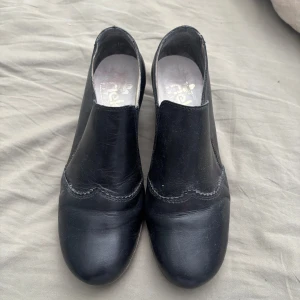 Svarta skinnloafers med klack från Rieker - Stilrena svarta loafers från Rieker i äkta skinn med låg klack och rund tå. Snygga sömdetaljer och elastiska partier på sidorna för enkel påtagning. Perfekta för dig som gillar klassisk och bekväm stil.