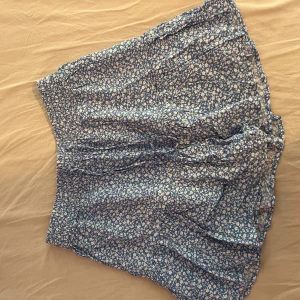 Blå blommig kortkjol med volang - Supersöt blå kortkjol med vitt blommönster och volangkant. Kjolen har resår i midjan och är luftig och lätt, perfekt för varma dagar. Den har även shorts under till. 