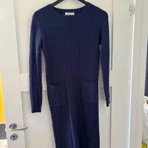 Mörkblå stickad långklänning Ebelieve - Mysig mörkblå långklänning från Ebelieve med kabelstickat mönster och två praktiska fickor framtill.