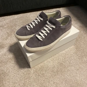 Common Projects Achilles Low Suede Mörkgrå - Snygga grå sneakers från Common Projects i mjuk mocka med vita snören och vit platt sula. Minimalistisk design med diskret guldfärgad numrering på hälen. Kommer med originalkartong, dustbag och extra skosnören. Nypris på 4000kr och slutsålda överallt.