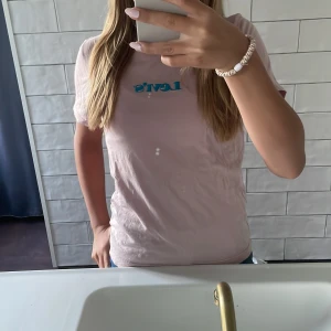 Ljusrosa Levi's t-shirt med blå logga - Ljusrosa t-shirt från Levi's med klassisk blå logga på bröstet. Modellen är rak med korta ärmar och rund halsringning. Perfekt basic-plagg med snygg och enkel stil, tillverkad i mjuk bomull.