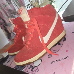 Röda Nike sneakers med kilklack - Snygga röda Nike sneakers i mocka med vit swoosh och beige sula. Skorna har snörning och en dold kilklack som ger extra höjd. Perfekta för dig som vill ha en sportig look med lite edge. Klassisk Nike-logga på plösen.