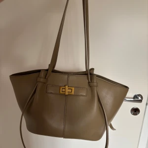 Beige axelväska från Gina Tricot - Stilren beige axelväska från Gina Tricot i slätt syntetmaterial. Väskan har långa axelband, snygg guldfärgad vridknäppning och rymligt innerfack med dragkedja. Perfekt för dig som vill ha en enkel men trendig väska med plats för allt du behöver.