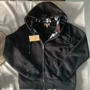 Svart Burberry hoodie med dragkedja, L men passar som M - Svart hoodie från Burberry med klassiskt rutigt foder i huvan och broderad logga på bröstet. Tröjan har hel dragkedja, snörning i huvan och ribbade muddar. 
