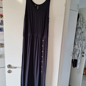 Svart långklänning från Rainbow - Svart långklänning från Rainbow med rund halsringning och resår i midjan. Klänningen har dekorativa knappar längs ena sidan och en slits nedtill. Tillverkad i mjuk viskosblandning med stretch för extra komfort.