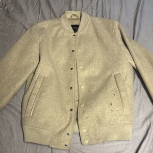 Beige bomberjacka från C&A Premium - Snygg beige bomberjacka från C&A Premium med klassisk ribbad krage och muddar. Jackan har knappar framtill, två fickor med läderdetaljer och är tillverkad i ullblandning. Perfekt för dig som gillar stilren och enkel design. smutsig men går nog bort i tvätten har inte testat
