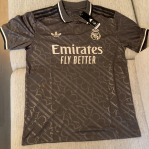 Real Madrid x Adidas grå fotbollströja - Snygg grå Real Madrid fotbollströja från Adidas med diskret mönster över hela tyget. Tröjan har korta ärmar, klassisk krage med vita detaljer och svarta Adidas-ränder på axlarna. Stort Emirates-tryck på bröstet och klubbmärke broderat. Perfekt för fans!