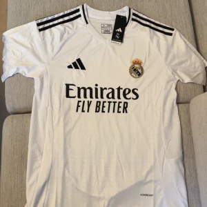 Real Madrid vit matchtröja Adidas - Säljer en officiell Real Madrid fotbollströja från Adidas i vitt med svarta detaljer och klubbmärke på bröstet. Tröjan har korta ärmar, svarta ränder på axlarna och är tillverkad i lätt, ventilerande material (AEROREADY). Perfekt för dig som älskar fotboll och Real.