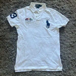 Vit pikétröja Polo Ralph Lauren M - Vit pikétröja från Polo Ralph Lauren i slim fit med klassisk krage och knäppning framtill. Stor broderad marinblå logga på bröstet och färgglada Yacht Club-detaljer. Kortärmad och tillverkad i mjuk bomull. Snygg och sportig stil med broderad siffra på ärmen.
