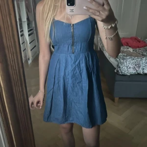 Blå jeansklänning med dragkedja - Supersöt blå jeansklänning med markerad midja och en snygg dragkedja framtill. Klänningen är kort och ärmlös, perfekt för varma dagar. Den har en lätt utställd kjol och sitter riktigt fint på kroppen. Enkel men ändå trendig look!