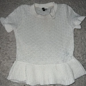 Vit stickad topp med volangkant - Supersöt vit stickad topp från H&M med korta ärmar och volangdetalj längst ner. Den har en rund halsringning och är perfekt för dig som gillar en feminin och trendig stil. Passar till både jeans och kjol för en fräsch look.