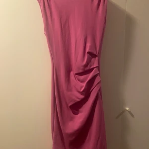 Rosa/lila klänning från Tiger of Sweden - Snygg rosa/lila klänning från Tiger of Sweden med draperad detalj på sidan. Modellen är ärmlös och har en figurnära passform som framhäver siluetten. Perfekt för dig som vill sticka ut med en färgstark look.