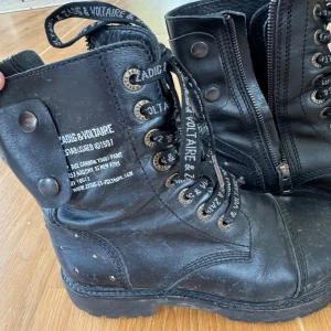 Svarta boots från Zadig & Voltaire - Tuffa svarta boots i skinn från Zadig & Voltaire med snörning, dragkedja på sidan och coola nitar i stjärnform baktill. Skorna har grov sula och logotyp på både snören och sidan. Perfekta för dig som gillar edgy stil.