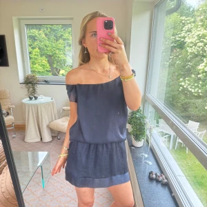 Mörkblå klänning i silke - Supersöt mörkblå kortklänning i silkesliknande material med omlott-detalj och offshoulder. Klänningen har en avslappnad passform och kan bäras både med axelband eller offshoulder. Perfekt för sommarens alla tillfällen! Passar S/M
