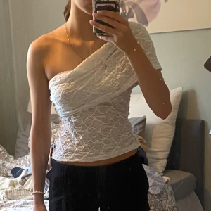 Vit spetsig oneshoulder topp - Säljer en vit topp i spets med oneshoulder-design. Toppen är figurnära och har ett snyggt omlott-utseende framtill. Den är ärmlös på ena sidan och täcker axeln på andra.