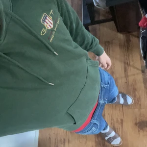 Grön hoodie från Gant med brodyr - Säljer en mörkgrön hoodie från Gant med broderad logga och sköld på bröstet. Tröjan har huva med snörning, känguruficka och ribbade muddar. Perfekt för dig som gillar sportig och avslappnad stil.