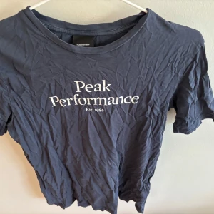 Mörkblå Peak Performance t-shirt - Snygg mörkblå t-shirt från Peak Performance med vit logga och text på bröstet. Klassisk rund halsringning och korta ärmar. Tillverkad i mjuk bomull som är skön att ha på sig. Perfekt för en avslappnad och sportig stil.