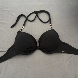 Svart bikiniöverdel push up - Snygg svart bikiniöverdel från Censored med push up och guldfärgade detaljer vid axelbanden. Bikinin har knyt i nacken och en stilren, enkel design som passar perfekt till sommaren. Materialet är mjukt och stretchigt för en bekväm passform. Säljer för den är för stor, aldrig använd.