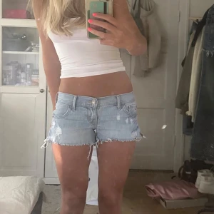 Ljusblå jeansshorts från Hollister - Snygga ljusblå jeansshorts från Hollister med låg midja och slitna detaljer. Råa, fransiga kanter och klassisk femficksmodell. Perfekta för sommaren och ger en avslappnad vibe. Ikonisk Hollister-logga på fickan och bak. Superbekväma och lätta att matcha. Midjemått:38cm, längden på shortsen är ca 25 cm