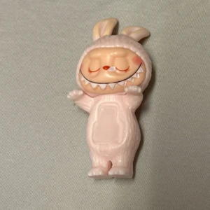 Mini Labubu Figur🌸 - Bilderna visar en liten labubu figur i rosa färg med ett stort leende och stängda ögon. Man kan klistra denna på telefonskalet. Köpte den i Cypern🌸