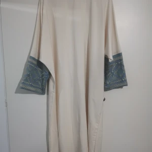 Långklänning/abaya  - Långklänning i ljus beige med vida ärmar som har blå tygdetaljer och broderade mönster i gult. Klänningen har en rak och lös passform, perfekt för dig som gillar unika detaljer och bohemisk stil.