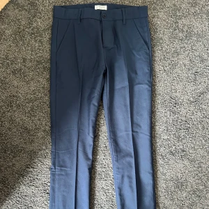 Kostymbyxor  - Stilrena och eleganta mörkblå kostymbyxor från Lindex – perfekta för både jobb och fest! Klassisk rak modell med pressveck, normal midja och bekväm passform. Tidlös design som enkelt matchas med kavaj, blus eller en enkel t-shirt för en mer avslappnad look. Använda endast ett fåtal gånger och i mycket gott skick – inga fläckar eller slitage.