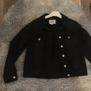 Svart jeansjacka från LAB Industries - Svart jeansjacka från LAB Industries med klassisk design, silverfärgade knappar och två bröstfickor med lock. Jackan har raka linjer och lång ärm, perfekt för dig som gillar en clean och tidlös look.