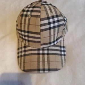  Keps från Burberry - Snygg keps från Burberry i klassiskt rutigt mönster med beige, svart, vitt och röda linjer. Justerbar rem baktill med metallspänne där Burberry är ingraverat. Perfekt accessoar för att lyfta din outfit med en lyxig touch. Säljer denna då jag köpte fel modell.
