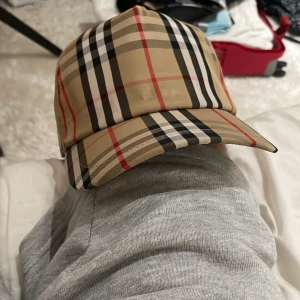 Rutig keps från Burberry i beige - Säljer en klassisk keps från Burberry med ikoniskt rutigt mönster i beige, svart, vitt och rött. Kepsen har böjd skärm och justerbar baksida med Burberry London England-patch. 