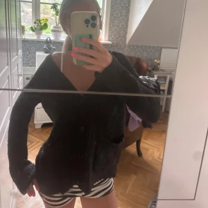 Svart kofta från Noa Noa - Mysig svart kofta från Noa Noa med v-ringning, stora knappar och två fickor framtill. Koftan är ribbstickad och har en loose passform med långa ärmar. Perfekt att slänga på sig när det är kyligt ute. 