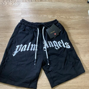Svarta Palm Angels shorts med tryck - Svarta shorts från Palm Angels med stort vitt logotyp-tryck över framsidan. De har elastisk midja med vita snören och avslappnad passform. Perfekta för en streetwear-look och riktigt sköna att bära under varma dagar. Storlek M.