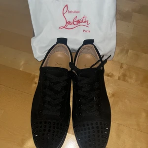 Christian Louboutin - Svarta sneakers från Christian Louboutin i mocka med ikoniska röda sulor och svarta nitar på tån. Skorna har snörning och en stilren, lyxig look som sticker ut. Perfekta för dig som vill ha något extra coolt och exklusivt i garderoben. Använda endast en gång och inga repor eller skador.