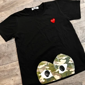  Comme des Garçons Play t-shirt - Svart t-shirt från Comme des Garçons Play med klassiskt rött hjärta på bröstet och stort camo-hjärta med ögon nertill. T-shirten är i mjuk bomull och har rund hals och korta ärmar. Perfekt för dig som bär storlek S och är 160-170cm lång!😁
