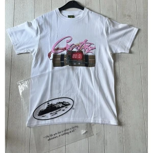 corteiz tröja - Vit t-shirt med färgstarkt grafiskt tryck framtill som föreställer en digital klocka och rosa detaljer. Klassisk passform och rund halsringning. Perfekt för dig som gillar unika prints och streetwear.