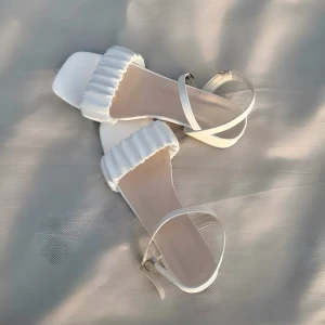 Vita sandaler med låg klack - Säljer ett par vita sandaler med låg klack, fyrkantig tå och volangdetalj över foten. Skorna har en smal rem runt vristen med spänne och platt sula. Perfekta för sommarens alla tillfällen. Endast använda 1 gång!