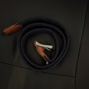 Svart flätat bälte med bruna skinn-detaljer - Säljer ett svart flätat bälte med bruna skinn-detaljer och silverfärgat spänne. Bältet har en stilren design och passar perfekt till jeans eller chinos. Enkel att justera för bästa passform. 90 cm