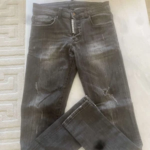 Svarta slim jeans från DSQUARED2 - Säljer ett par svarta slim fit jeans från DSQUARED2 med slitningar och diskreta detaljer. Jeansen har klassisk femficksdesign, knappgylf och tydlig logga på både framsidan och insidan. Perfekta för dig som gillar en modern och edgy stil.