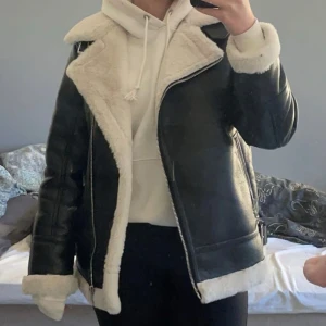 Svart skinnjacka med foder - Säljer en svart skinnjacka med vitt teddyfoder och dragkedja. Jackan har bred krage, långa ärmar och synliga sömmar. Perfekt för dig som vill ha en cool och varm look till hösten eller vintern.