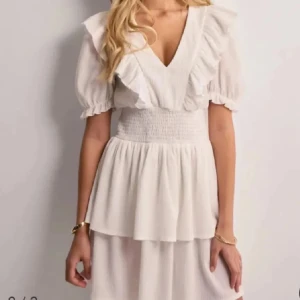 Vit volangklänning från Gina Tricot - Superfin vit klänning från Gina Tricot med v-ringning, volangdetaljer och kort ärm. Smockad midja ger en snygg passform och kjolen har ett lätt fall. Perfekt för sommarens alla tillfällen! 