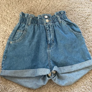 Blå jeansshorts med resårmidja från Lindex - Säljer ett par blå jeansshorts från Lindex med hög resårmidja och uppvikta benslut. Shortsen har två framfickor, två bakfickor och bälteshällor. Perfekta för varma dagar och enkel att matcha med olika toppar.
