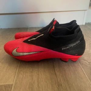 Nike Phantom Vision ACC  - Säljer ett par Nike Phantom Vision fotbollsskor i rött och svart med silverdetaljer. Skorna har en strumpliknande passform och stora silvriga Nike-loggor på sidorna. Perfekta för dig som vill ha kontroll på planen.
