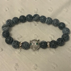 Svart pärlarmband med leopardhuvud - Coolt armband med svarta pärlor och ett glittrigt leopardhuvud i silver med gröna ögon. Mellan pärlorna sitter små detaljer med strass. Perfekt accessoar för att sticka ut lite extra.